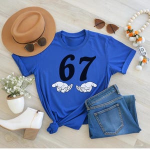 May include: A royal blue t-shirt with the number 67 in black, and two cartoon hands. The shirt is paired with blue jeans, a tan hat, sunglasses, white boots, and a beaded necklace with orange accents.
