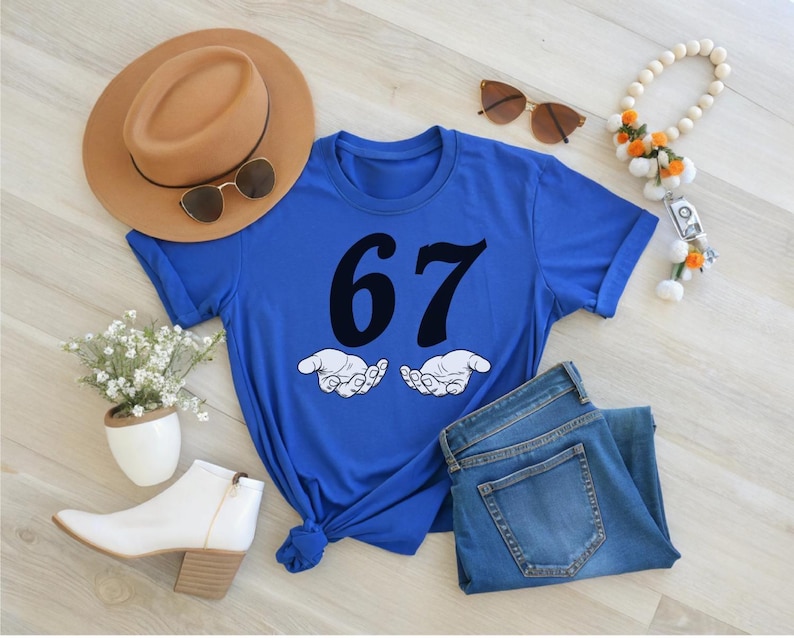 May include: A royal blue t-shirt with the number 67 in black, and two cartoon hands. The shirt is paired with blue jeans, a tan hat, sunglasses, white boots, and a beaded necklace with orange accents.