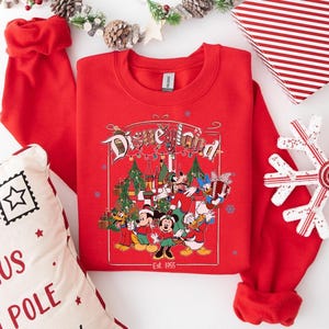 K&ouml;nnte beinhalten: Ein rotes Sweatshirt mit Rundhalsausschnitt und einem Disneyland-Motiv mit Micky Maus, Minnie Maus, Donald Duck und Goofy. Die Grafik enth&auml;lt Weihnachtsb&auml;ume und den Text "Est. 1955". Ein dekorativer Schneeflocke, eine Geschenkbox und ein Kissen sind ebenfalls sichtbar.