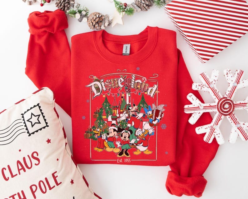 K&ouml;nnte beinhalten: Ein rotes Sweatshirt mit Rundhalsausschnitt und einem Disneyland-Motiv mit Micky Maus, Minnie Maus, Donald Duck und Goofy. Die Grafik enth&auml;lt Weihnachtsb&auml;ume und den Text "Est. 1955". Ein dekorativer Schneeflocke, eine Geschenkbox und ein Kissen sind ebenfalls sichtbar.