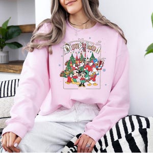 K&ouml;nnte beinhalten: Hellrosa Sweatshirt mit Disneyland-Grafik. Die Grafik zeigt Micky, Minnie, Donald und Goofy vor einem Weihnachtsbaum und einem Schloss. Das Wort "Disneyland" ist oben in einem Bogen angeordnet.
