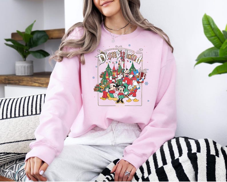 K&ouml;nnte beinhalten: Hellrosa Sweatshirt mit Disneyland-Grafik. Die Grafik zeigt Micky, Minnie, Donald und Goofy vor einem Weihnachtsbaum und einem Schloss. Das Wort "Disneyland" ist oben in einem Bogen angeordnet.