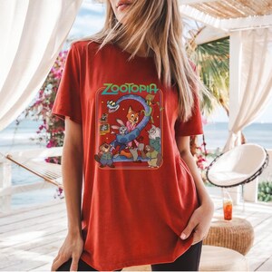 May include: Red t-shirt featuring the word "Zootopia" in a colorful, retro-style design. The graphic includes cartoon characters from the movie. The shirt is made of a soft material.