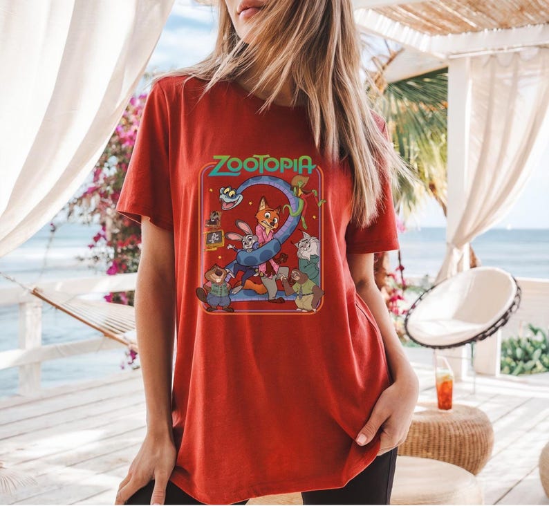 May include: Red t-shirt featuring the word "Zootopia" in a colorful, retro-style design. The graphic includes cartoon characters from the movie. The shirt is made of a soft material.