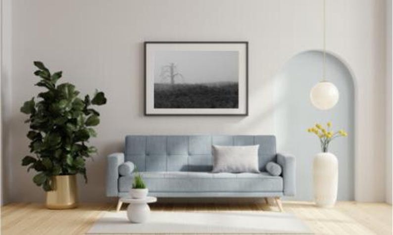 May include: A living room scene featuring a light blue sofa with a white pillow, a framed black and white landscape print, and a large potted plant. A white vase holds yellow flowers, and a pendant light hangs from the ceiling.