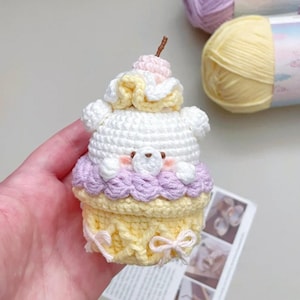 May include: A handmade crochet cupcake-shaped plush toy in white, yellow, and lavender. The toy features a bear-like face, a cherry on top, and a decorative bow. The yarn is visible in the background.
