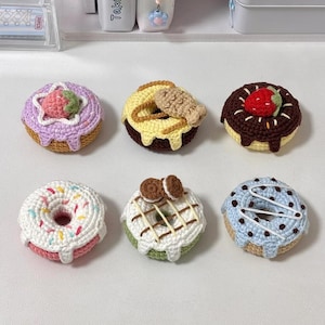 May include: A collection of six crocheted donuts in various colors and designs. Each donut features unique toppings, including sprinkles, chocolate, and fruit. The donuts are arranged on a white surface, showcasing the handmade details and vibrant colors.