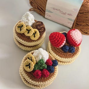 Könnte beinhalten: Drei gehäkelte Pfannkuchen mit verschiedenen Belägen, darunter Bananen, Erdbeeren, Blaubeeren und Schlagsahne. Die Pfannkuchen sind gestapelt und haben eine beige-braune Farbgebung. Im Hintergrund ist ein Knäuel brauner Wolle zu sehen.