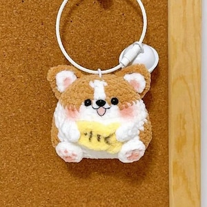 May include: A plush corgi keychain with brown, white, and pink fur, holding a yellow sign with Japanese text. The keychain has a white metal ring and is attached to a corkboard within a wooden frame.