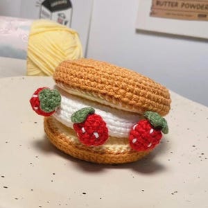 May include: Crocheted dessert resembling a strawberry shortcake. The top and bottom layers are tan, with a white filling. Red strawberries with green leaves are attached to the sides. The dessert is on a white plate.