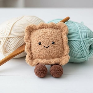May include: A handmade, plush, smiling ravioli toy with brown legs, a wooden crochet hook, and two yarn balls. The ravioli is light brown with a textured surface. One yarn ball is cream-colored, and the other is light blue.