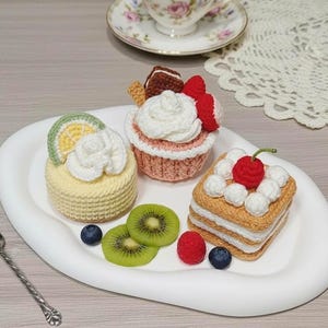 Op de afbeelding: Een witte serveerschaal met drie gehaakte desserts: een gele cake met een citroenschijfje, een roze cupcake met een aardbei en een gelaagde cake met een kers. Kiwischijfjes, bosbessen en frambozen liggen ook op de schaal.