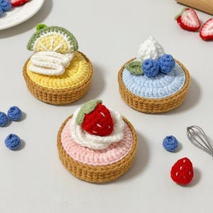 Op de afbeelding: Drie gehaakte miniatuartaartjes met fruit toppings, waaronder een citroenschijfje, blauwe bessen en een aardbei. Extra gehaakte blauwe bessen en aardbeien zijn verspreid rond de taartjes. Een deegroller en garde zijn ook aanwezig.