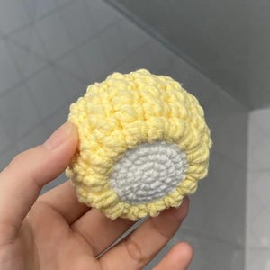 Op de afbeelding: Een handgemaakte, gele gehaakte bal met een wit midden. De bal wordt in een hand gehouden en toont de textuur en het vakmanschap van het garen. De haaksteken creëren een getextureerde, bolvormige vorm.