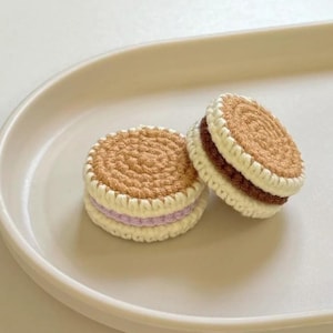 May include: Two handmade crochet coasters shaped like cookies. The coasters feature a light brown top and bottom with a white border and a contrasting filling of either purple or brown. They are displayed on a white oval plate.
