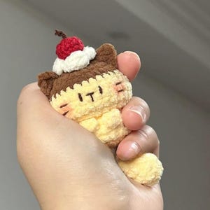 May include: A crocheted cat-shaped plush toy in yellow, brown, and white, topped with a red cherry. The toy has the letters "IT" embroidered on its face. The cat's ears and top are brown, with a white cream-like layer and a red cherry on top.