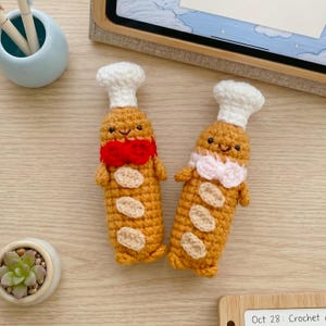 May include: Two crocheted baguette figures with smiling faces, each wearing a white chef hat. One baguette features a red bow tie, while the other has a light pink bow tie. Both figures have small tan oval patches on their bodies and tiny arms.