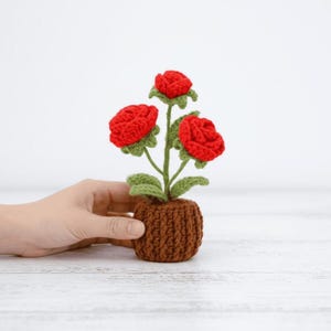 May include: A crocheted flower arrangement with three red roses, green leaves and stems, in a brown crocheted pot. The craftwork displays detailed stitching and a realistic floral design, perfect for home decor.
