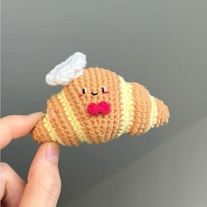 Puede incluir: Croissant de ganchillo con gorro de chef y pajarita. La pastelería es de color marrón con rayas amarillas, una pajarita roja y un gorro de chef blanco. El croissant tiene una cara sonriente con ojos negros y boca negra.