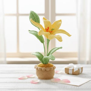 May include: A handcrafted crochet lily flower in a small pot. The flower has yellow petals with orange accents, green leaves, and a green stem. The pot is tan with a scalloped edge. A small gift box with a white ribbon and a blank card are in the background.