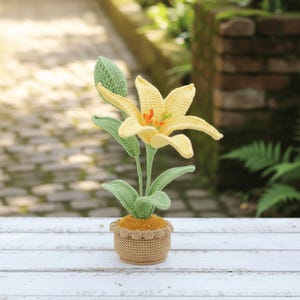 May include: A handmade crochet lily flower in a small pot. The flower has yellow petals with orange accents, green leaves, and a green stem. The pot is tan with a decorative edge, sitting on a white wooden surface.