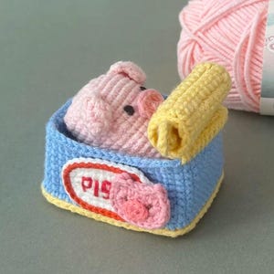 May include: A crocheted amigurumi pig inside a blue rectangular container with a yellow rolled item. The container features a white oval label with a red border and the red text PIG, accompanied by a small pink crocheted pig face appliqué.
