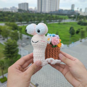 Puede incluir: Un juguete de caracol hecho a mano con ganchillo, con ojos grandes con bordes blancos y una cara sonriente. La concha del caracol es una cesta marrón llena de flores de ganchillo de colores. El caracol se sostiene frente a un fondo borroso al aire libre.