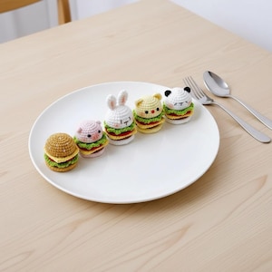 Op de afbeelding: Vijf kleine, ronde, gehaakte amigurumi-figuurtjes in de vorm van miniatuurhamburgers, elk met een ander dierenhoofd, waaronder een gewoon broodje, een varken, een konijn, een kat en een panda, allemaal op een rij op een groot wit bord.