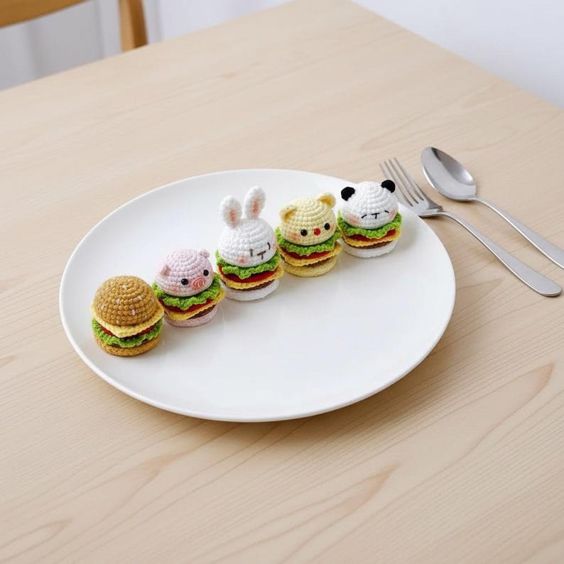 Op de afbeelding: Vijf kleine, ronde, gehaakte amigurumi-figuurtjes in de vorm van miniatuurhamburgers, elk met een ander dierenhoofd, waaronder een gewoon broodje, een varken, een konijn, een kat en een panda, allemaal op een rij op een groot wit bord.