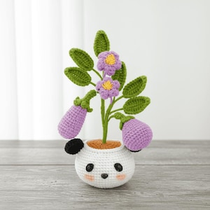 Puede incluir: Una planta de ganchillo en una maceta blanca con forma de panda. La planta presenta flores moradas, hojas verdes y dos berenjenas moradas. La maceta tiene ojos negros, una oreja negra y mejillas rosadas. La planta está sobre una superficie de madera.