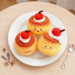 Może przedstawiać: Trzy okrągłe, szydełkowane desery amigurumi przypominające budynie, z żółtymi podstawami, brązowymi wierzchami, białą bitą śmietaną i czerwonymi wiśniami. Każdy element ma unikalną haftowaną twarz z małymi oczami i ustami na żółtym korpusie.