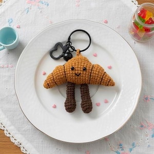 Pode incluir: Um chaveiro de crochê em forma de croissant sorridente com pernas marrons. O croissant é marrom claro com listras marrons escuras. Possui um fecho e um anel pretos. O fundo inclui um prato branco e uma xícara azul pequena.