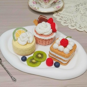 Op de afbeelding: Drie gehaakte dessertreplica's op een witte ovale schaal. Een gele ronde cake met een schijfje limoen en room, een perzikkleurige cupcake met room en bessen, en een vierkante gelaagde cake met witte room en een rode kers bovenop.