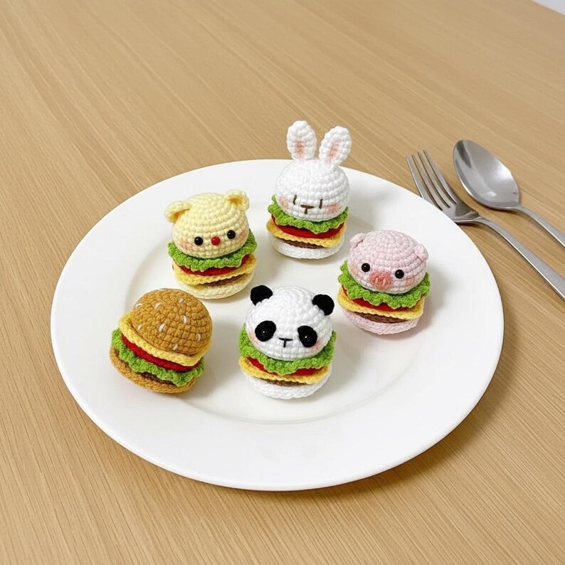 Op de afbeelding: Vijf kleine, gehaakte amigurumi-figuurtjes in de vorm van miniatuurhamburgers, elk bekroond met een andere dierenkop: een bruin broodje, een wit konijn, een gele beer, een zwart-witte panda en een lichtroze varken, op een wit bord.