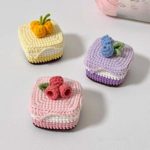 May include: Three square crochet cakes featuring layered designs. One is yellow with orange fruit, one is lavender with blue berries, and one is pink with red berries. Each cake has a white middle layer and green crocheted leaves.
