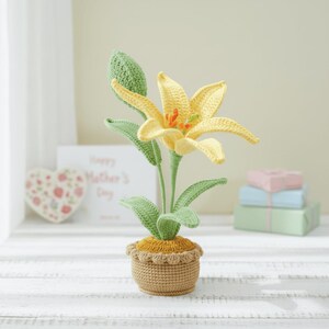 May include: A handcrafted crochet lily flower in a small pot. The flower has yellow petals with orange accents, green leaves, and a brown pot. The pot has a scalloped edge. The lily is displayed on a white wooden surface.