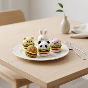 Op de afbeelding: Een verzameling van vijf kleine, gehaakte knuffels in de vorm van hamburgers, elk bekroond met een andere dierenkop, waaronder een beer, konijn, panda en varken. De kleurrijke garencreaties zijn gerangschikt op een wit bord met bestek.