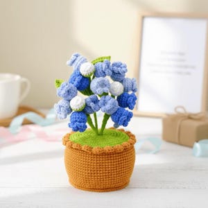 Pode incluir: Um arranjo de flores de crochê feito à mão em um pequeno vaso laranja. As flores são azuis, azul claro e brancas, com folhas verdes. O vaso tem uma borda ondulada. Um item decorativo.
