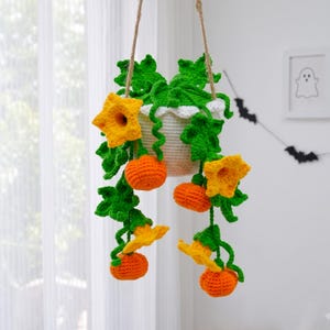 May include: A hanging crochet plant featuring a white pot, vibrant green leaves, bright yellow trumpet-shaped flowers, and small orange pumpkins. The entire piece is suspended by a thin brown cord, showcasing intricate yarn textures and colors.