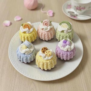 以下が含まれることがあります： 白い皿の上に並べられた6つのカップケーキ型のかぎ針編みオーナメント。黄色、ピンク、緑、青、紫などのパステルカラーのテクスチャーのある土台に、白いかぎ針編みのアイシングと小さなフルーツやクッキーのアクセントが施されています。