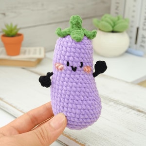 May include: A handmade, crocheted eggplant plushie in shades of purple, green, and black. The eggplant has a smiling face with rosy cheeks and small black arms. The plushie is held in a person's hand.
