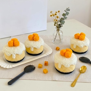 Könnte beinhalten: Drei gehäkelte Kuchen mit hellgelben Basen, weißem Zuckerguss und jeweils drei kleinen orangefarbenen Kugeln. Die Kuchen sind mit einer schwarzen Plastikgabel, einem goldenen Löffel und kleinen gehäkelten orangefarbenen Würfeln arrangiert.