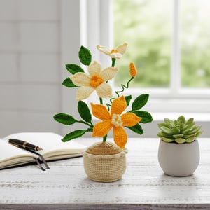 May include: A handcrafted crochet flower arrangement featuring orange and cream-colored blossoms with green leaves, presented in a beige crochet pot. A small succulent in a white pot sits nearby. A notebook and pen are also visible.