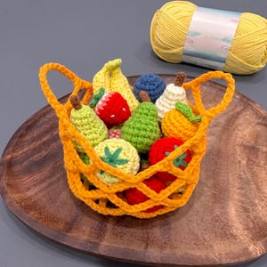 Peut inclure: Un panier orange vif au crochet rempli de divers petits fruits et légumes crochetés colorés, dont une banane, une fraise, une poire et une tomate. Une pelote de laine jaune est posée à proximité, complétant cet ensemble d'objets textiles.