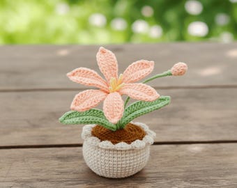 May include: A crocheted flower in a small pot. The flower has peach-colored petals and a yellow center. Green leaves and a bud accompany the flower. The pot is white with a scalloped edge and sits on a wooden surface.