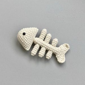 May include: A cream-colored crochet fish skeleton ornament featuring a textured yarn construction. The design includes a rounded head section with a single small black bead eye and a central spine with four pairs of protruding ribs and a tail.