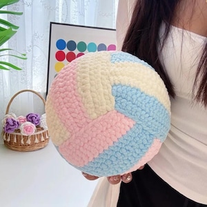 Op de afbeelding: Een ronde, veelkleurige bal met een getextureerd oppervlak. De bal heeft secties van lichtblauw, roze en crèmekleurig garen. Op de achtergrond staat een klein mandje met decoratieve bloemen.