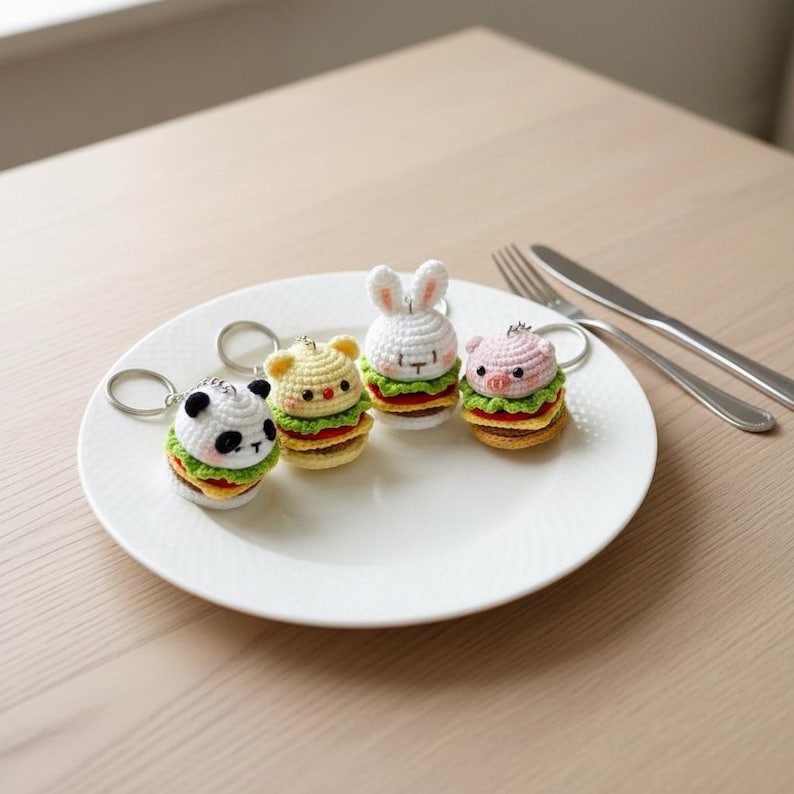 Op de afbeelding: Vier gehaakte sleutelhangers in de vorm van miniatuurhamburgers met dierenkoppen, waaronder een panda, een beer, een konijn en een varken. Elke sleutelhanger is voorzien van een zilverkleurige metalen ring aan de bovenkant van het figuurtje.