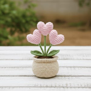 May include: A crocheted plant featuring three pink heart-shaped flowers, green stems, and leaves, presented in a beige pot. The pot contains brown material, with a blurred green backdrop.
