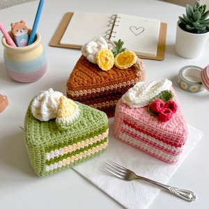 Peut inclure: Trois parts de gâteau triangulaires au crochet en vert, marron et rose. La part verte a un glaçage blanc et un quartier jaune. La part marron présente des fleurs blanches et des fruits jaunes. La part rose a un glaçage blanc et des baies rouges.